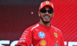 Smiling man in a red Ferrari cap and shirt with sponsor logos, wearing sunglasses against a red background.