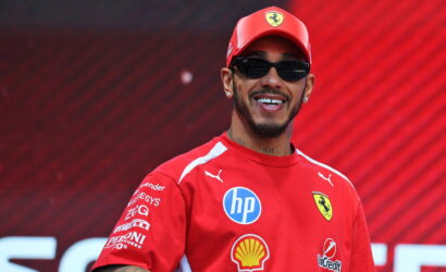 Smiling man in a red Ferrari cap and shirt with sponsor logos, wearing sunglasses against a red background.