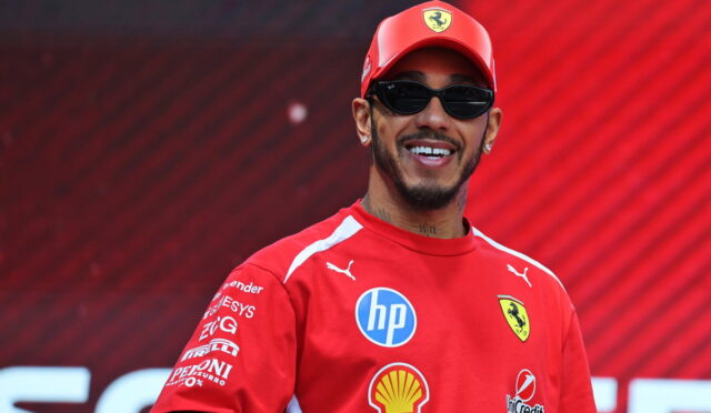 Smiling man in a red Ferrari cap and shirt with sponsor logos, wearing sunglasses against a red background.