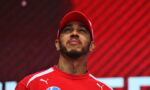 Portrait of a man in a red cap and red athletic shirt looking upward, neck tattoos visible against a red diagonal background.