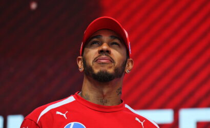 Athlete in a red racing jersey and cap, looking upward with a red patterned background behind him.