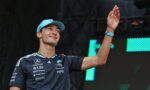 Young man in a blue cap waves on stage, wearing a dark sponsor-logo jersey and a blue wristband.