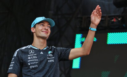 Young man in a blue cap waves on stage, wearing a dark sponsor-logo jersey and a blue wristband.