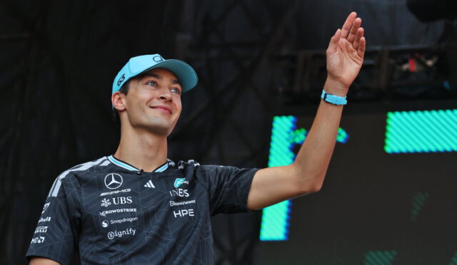 Young man in a blue cap waves on stage, wearing a dark sponsor-logo jersey and a blue wristband.