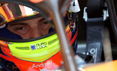 Close-up of a race car driver wearing a bright neon-yellow helmet with Android text, seen through the helmet visor inside a cockpit.