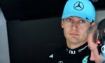 Close-up of a Formula 1 driver in a bright blue Mercedes cap, black racing suit with Petronas logos, looking to the left.