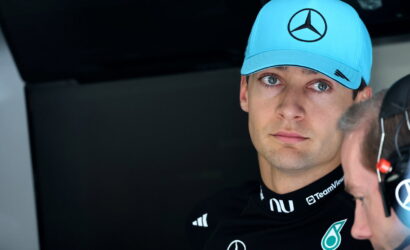 Close-up of a Formula 1 driver in a bright blue Mercedes cap, black racing suit with Petronas logos, looking to the left.