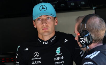 Formula 1 driver in a black Mercedes suit and teal cap listens to a team engineer wearing a headset in the garage