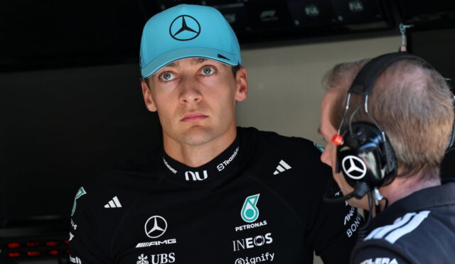 Formula 1 driver in a black Mercedes suit and teal cap listens to a team engineer wearing a headset in the garage