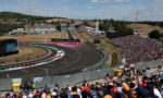 Crowded motorsport grandstand overlooking a winding race track on a sunny day, with a race car on the curve.