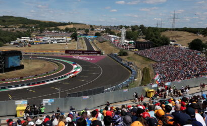 Crowded motorsport grandstand overlooking a winding race track on a sunny day, with a race car on the curve.
