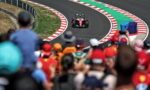 Red Formula 1 race car on a winding track, seen from the crowded stands with red-and-white striped curbs and barriers along the curve.