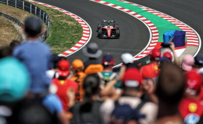 Red Formula 1 race car on a winding track, seen from the crowded stands with red-and-white striped curbs and barriers along the curve.