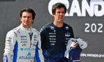 Two race drivers in white and dark blue suits stand together on a podium after a race event, looking off to the side.