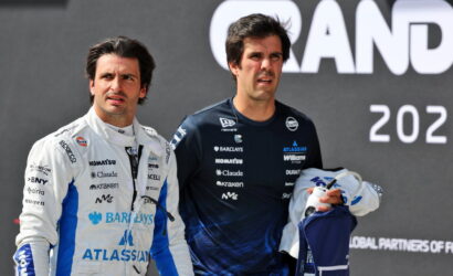 Two race drivers in white and dark blue suits stand together on a podium after a race event, looking off to the side.