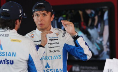 Race car driver in a white-blue branded suit adjusts his hands, while a teammate in the foreground faces away.