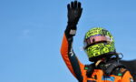 Racing driver in an orange suit raises a gloved hand against a clear blue sky.