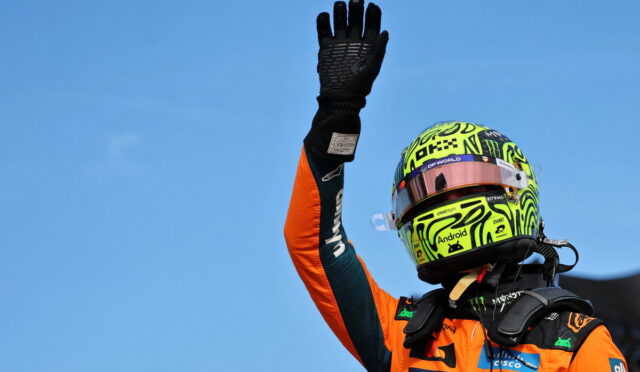 Racing driver in an orange suit raises a gloved hand against a clear blue sky.
