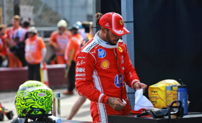 Ferrari driver in a red racing suit stands at a pit wall, handling a white towel with a yellow helmet and gear nearby.