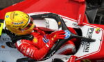 Ferrari Formula 1 driver in a red racing suit gripping the steering wheel inside a red-and-white F1 car, wearing a yellow helmet with sponsor logos.