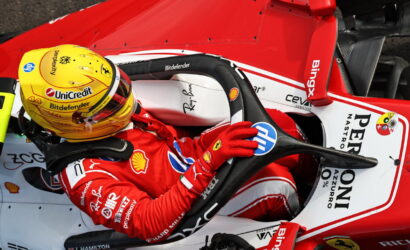 Ferrari Formula 1 driver in a red racing suit gripping the steering wheel inside a red-and-white F1 car, wearing a yellow helmet with sponsor logos.