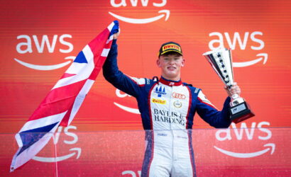 Race car driver on a podium holds a tall trophy in one hand and a British flag in the other, against a red AWS backdrop.