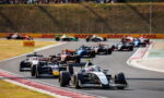 Close-packed open-wheel race cars navigate a curving track, leading car #16 at the front with spectators in the hillside stands.