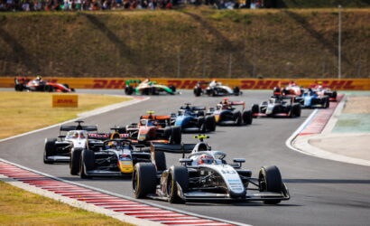 Close-packed open-wheel race cars navigate a curving track, leading car #16 at the front with spectators in the hillside stands.