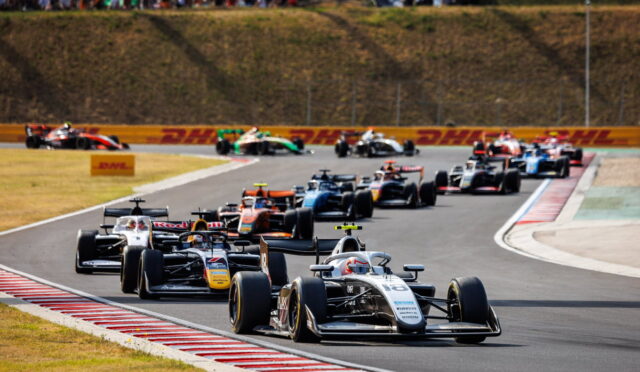 Close-packed open-wheel race cars navigate a curving track, leading car #16 at the front with spectators in the hillside stands.