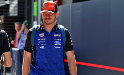 Male race team member wearing a blue Oracle Red Bull Racing polo and orange cap, standing with a backpack in a paddock area.