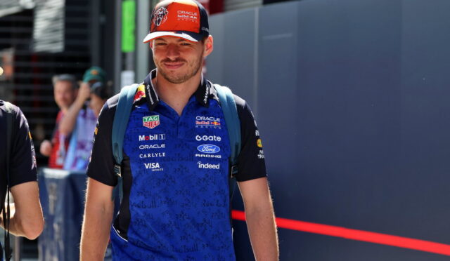 Male race team member wearing a blue Oracle Red Bull Racing polo and orange cap, standing with a backpack in a paddock area.