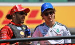 Two Formula 1 drivers wearing sunglasses chat on the track, one in red Ferrari gear with a red cap, the other in white Mercedes gear with a blue cap.