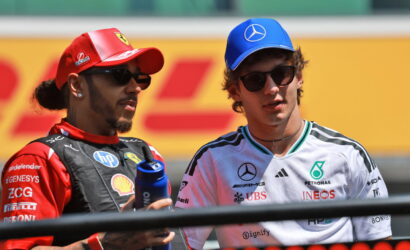Two Formula 1 drivers wearing sunglasses chat on the track, one in red Ferrari gear with a red cap, the other in white Mercedes gear with a blue cap.