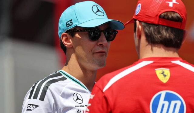 Two race team members face each other in conversation: a man in a light-blue Mercedes cap and sunglasses wearing a white racing shirt with sponsor logos, and a red Ferrari-clad teammate seen from behind with an HP logo on the back.