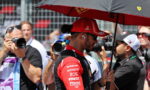 Race car driver in a red suit and cap stands under a black umbrella while photographers surround him at a race event.