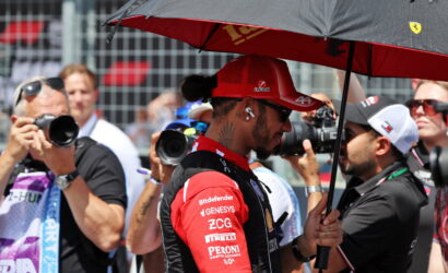 Race car driver in a red suit and cap stands under a black umbrella while photographers surround him at a race event.