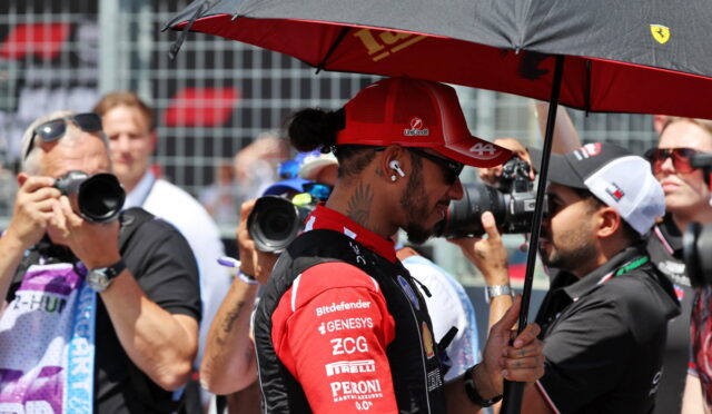 Race car driver in a red suit and cap stands under a black umbrella while photographers surround him at a race event.