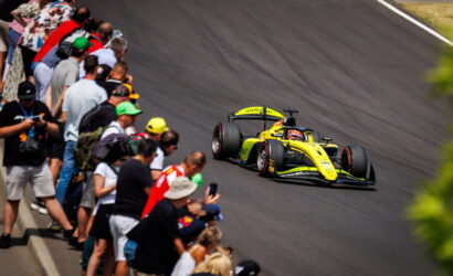Yellow open-wheel race car speeds along a curve as a crowd of spectators lines the track on the left, watching closely.
