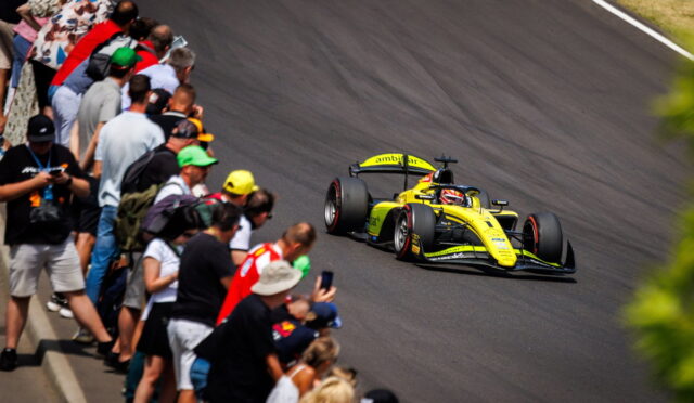 Yellow open-wheel race car speeds along a curve as a crowd of spectators lines the track on the left, watching closely.