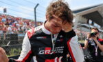 Race car driver in GR Toyota Gazoo Racing suit with hand on his forehead, crowd and a photographer in the background, suggesting a post-race moment.