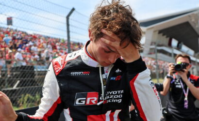 Race car driver in GR Toyota Gazoo Racing suit with hand on his forehead, crowd and a photographer in the background, suggesting a post-race moment.