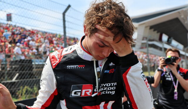 Race car driver in GR Toyota Gazoo Racing suit with hand on his forehead, crowd and a photographer in the background, suggesting a post-race moment.