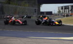 Formula 1 race cars cornering on a track, red and blue RB livery cars leading a pack near a barrier fence and grandstand.