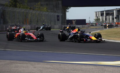 Formula 1 race cars cornering on a track, red and blue RB livery cars leading a pack near a barrier fence and grandstand.