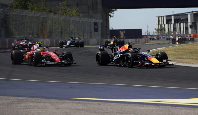Formula 1 race cars cornering on a track, red and blue RB livery cars leading a pack near a barrier fence and grandstand.