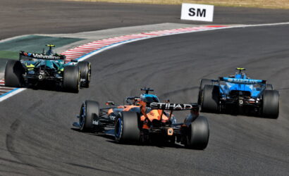 Three Formula 1 cars navigate a tight curving track, with orange Etihad car in the lead and blue Barclays car trailing on the right while a black Valvoline car follows on the left near red-and-white curb stripes.