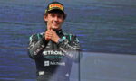 Formula 1 driver in a racing suit giving a thumbs-up on stage, wearing a Pirelli cap and Petronas-Microsoft livery