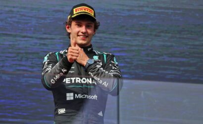 Formula 1 driver in a racing suit giving a thumbs-up on stage, wearing a Pirelli cap and Petronas-Microsoft livery