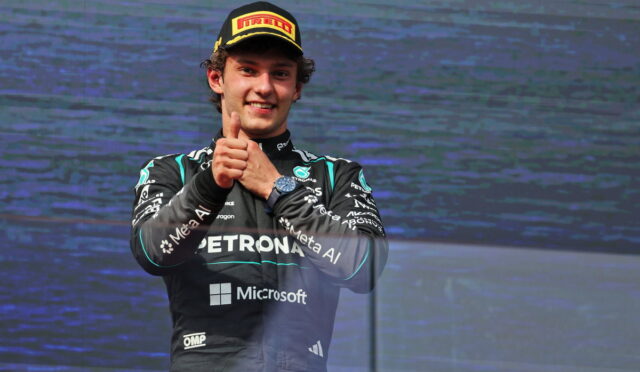 Formula 1 driver in a racing suit giving a thumbs-up on stage, wearing a Pirelli cap and Petronas-Microsoft livery