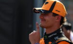 Portrait of a male race driver in an orange cap and racing suit, with his finger to his lips in a shushing gesture.